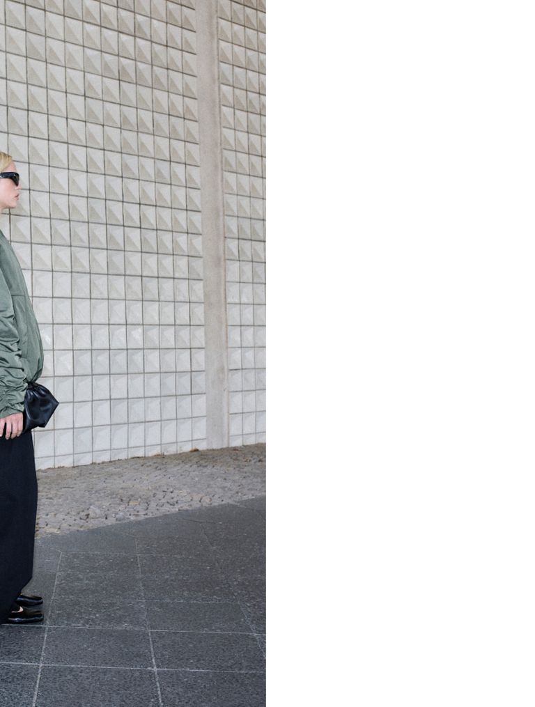 Person in a green jacket and sunglasses stands by a textured wall, holding a black bag, with a spacious, tiled environment in the background.
