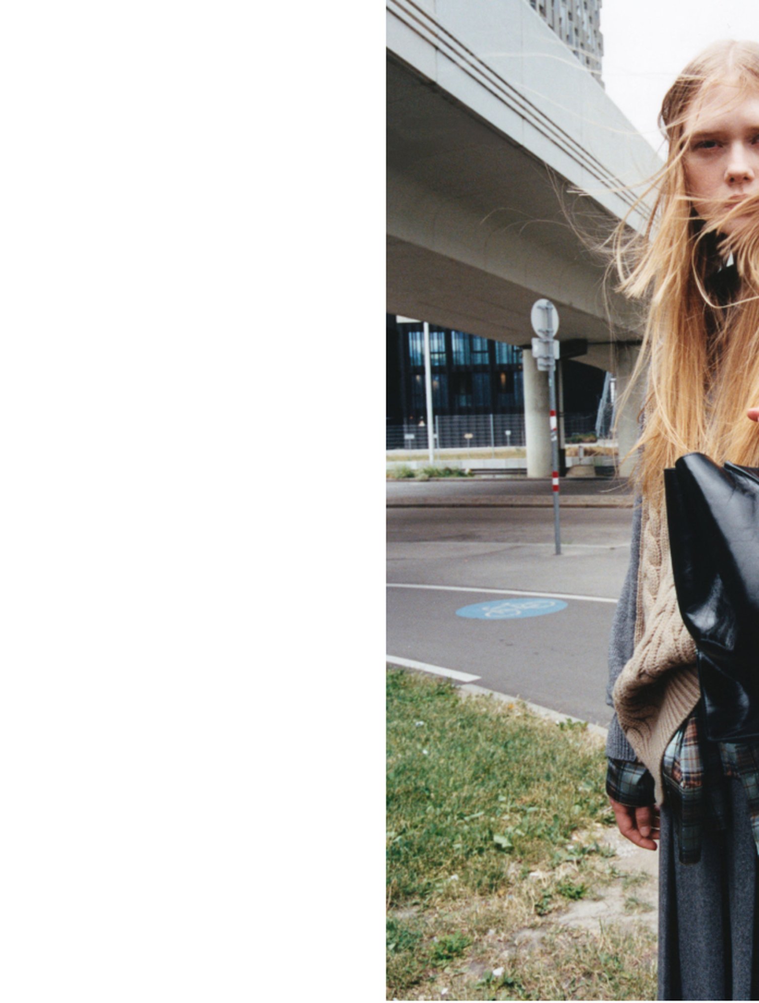 Person with long hair stands near a road under a bridge, holding a black bag. Wind blows their hair across their face.