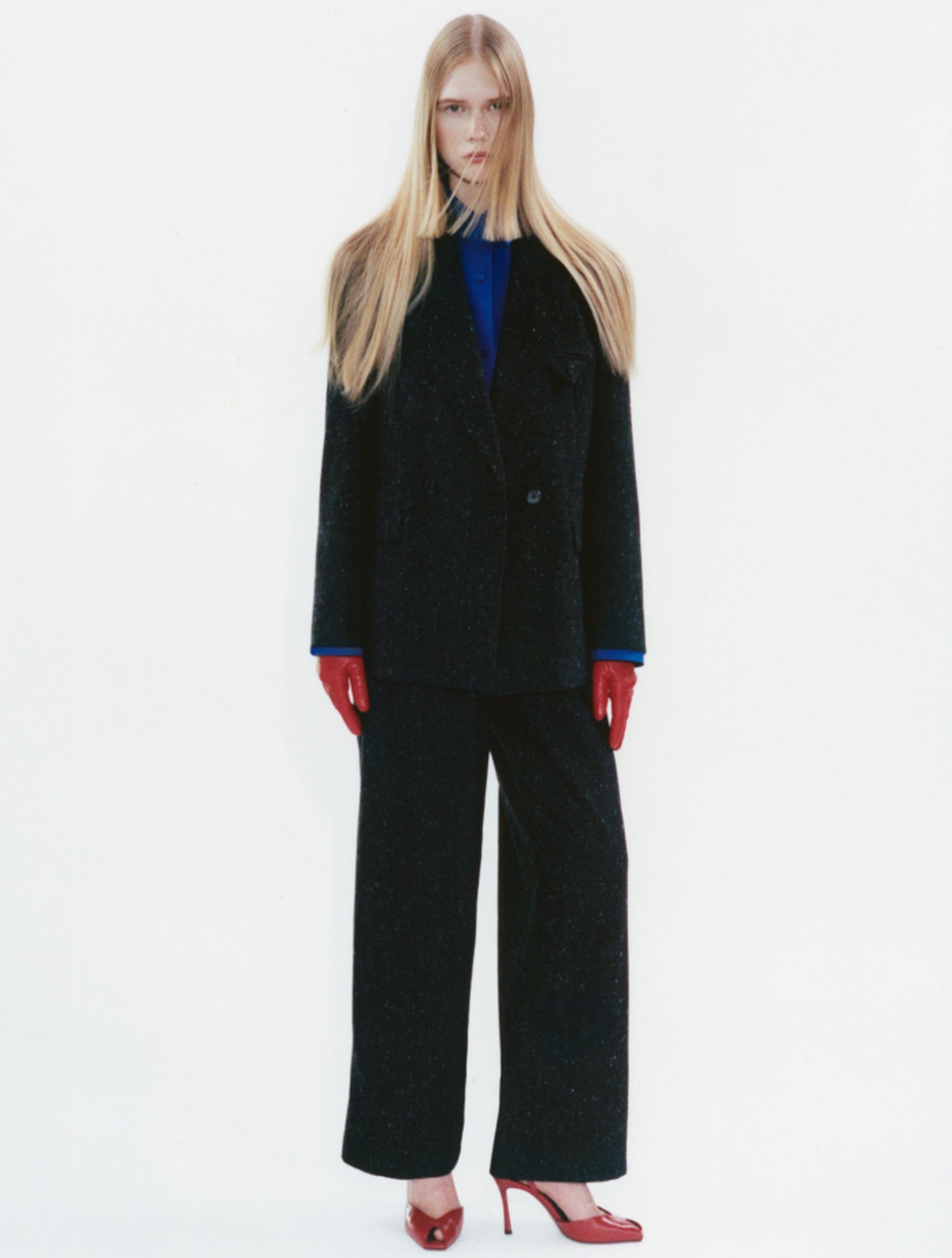 Person with long hair in a dark oversized suit, blue shirt, red gloves, and red heels, standing against a plain white background.