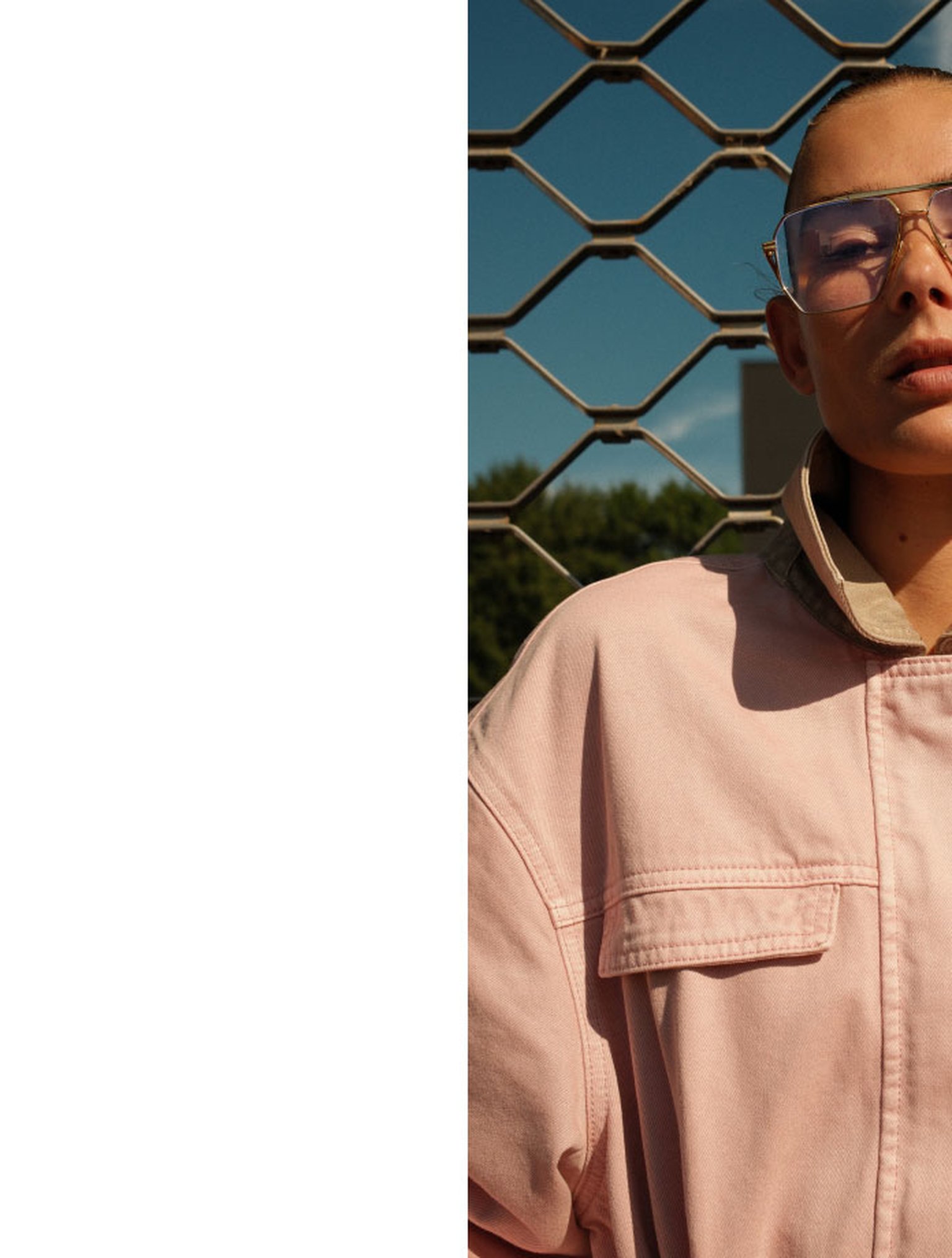 Person wearing a pink jacket and glasses stands in front of a chain-link fence with a clear blue sky in the background.