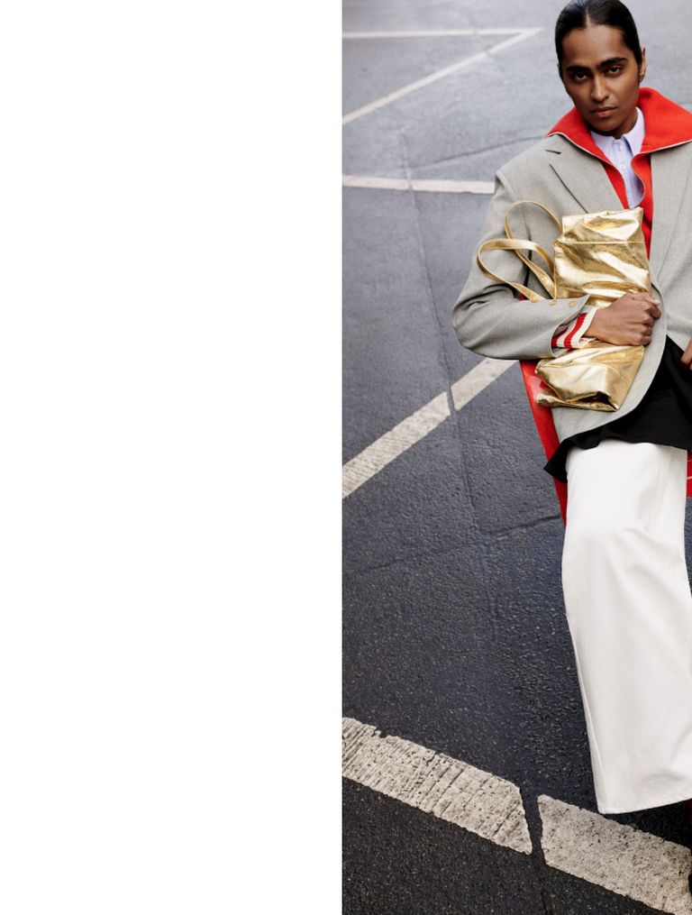Fashion model in a gray coat with red lapels, holding a golden bag, poses confidently on a crosswalk.