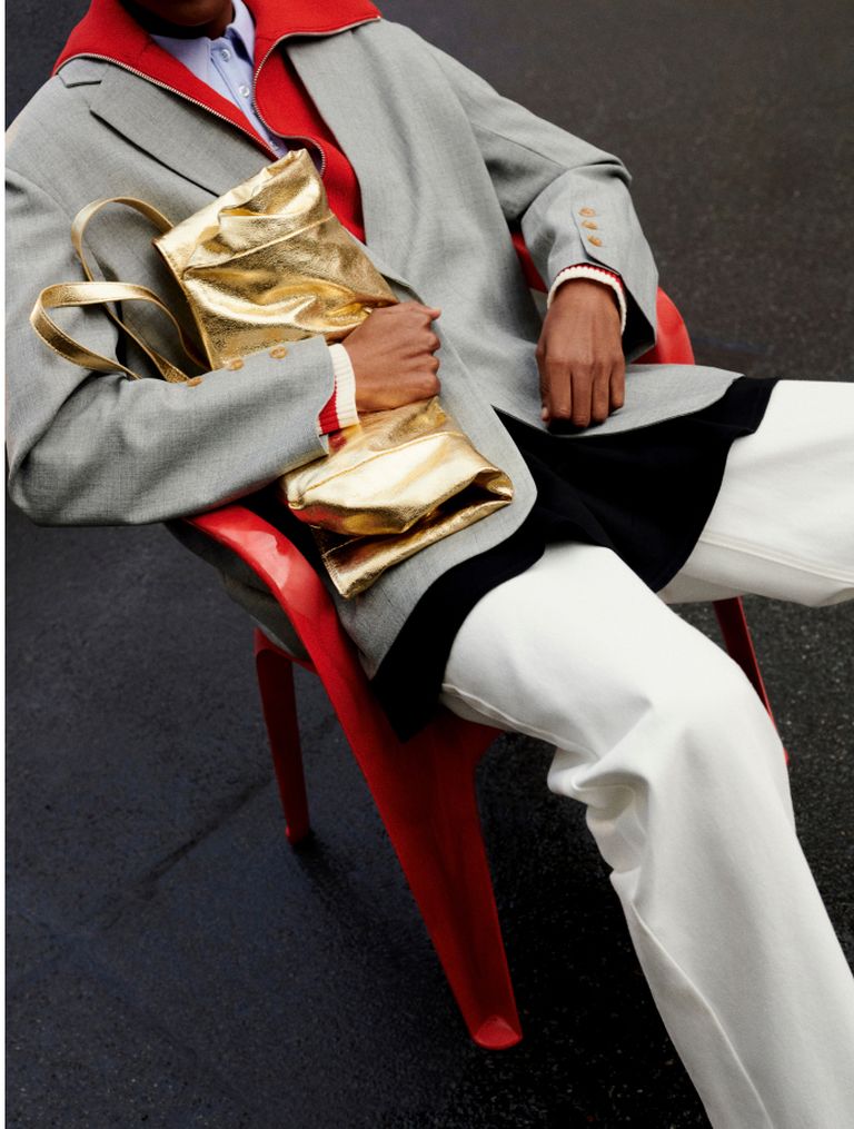 Person in a gray coat with red collar, holding a shiny gold bag, sits on a red chair, wearing white pants against a dark background.