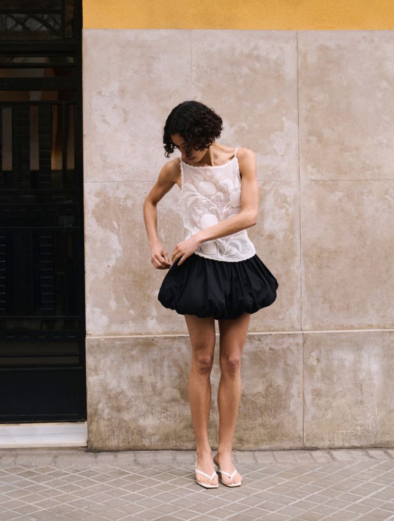 Person in a white lace top and black skirt stands against a beige wall, adjusting their outfit, wearing flip-flops.