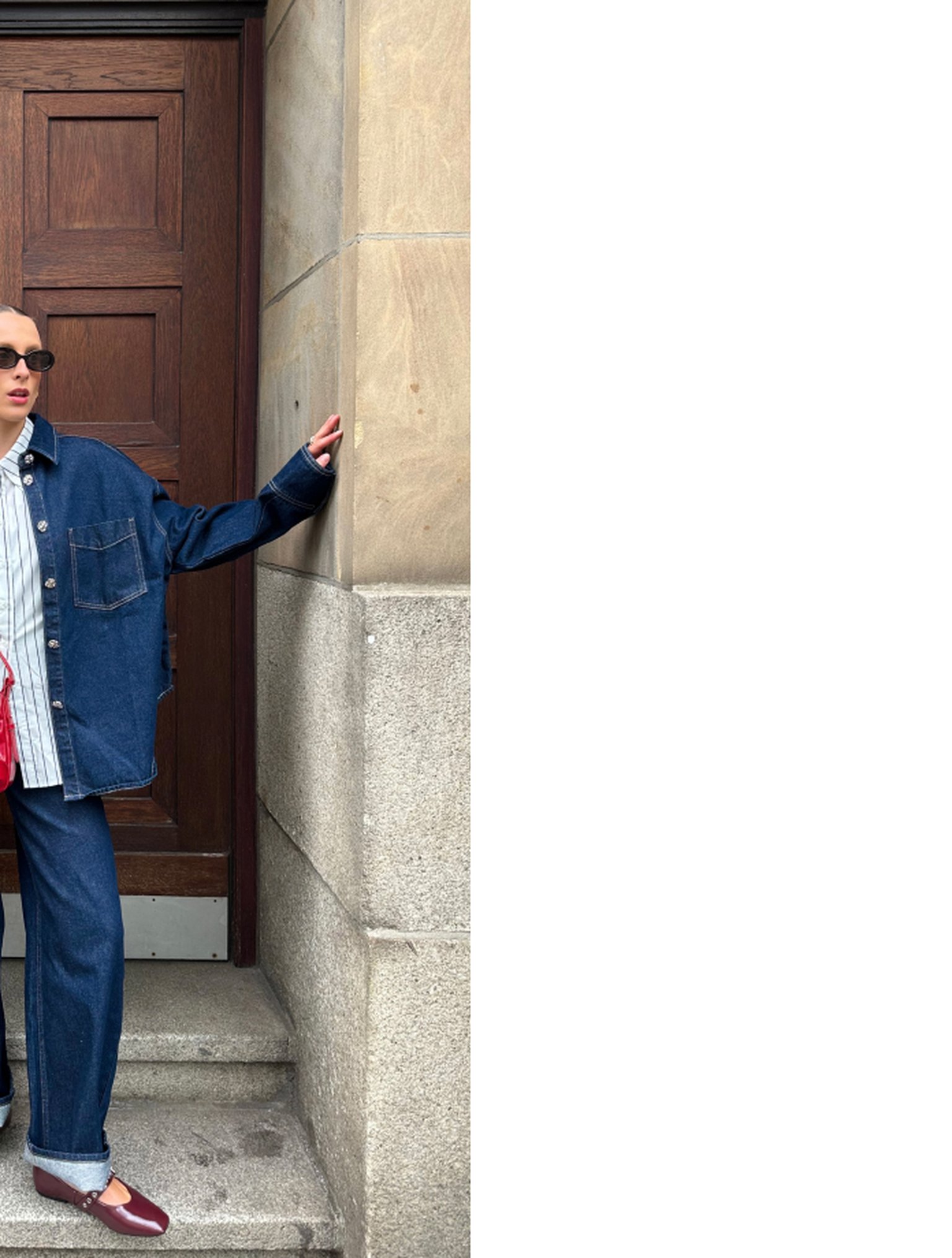 Person in a denim jacket and jeans, wearing sunglasses, leans against a stone wall near a wooden door, with one hand on the wall.