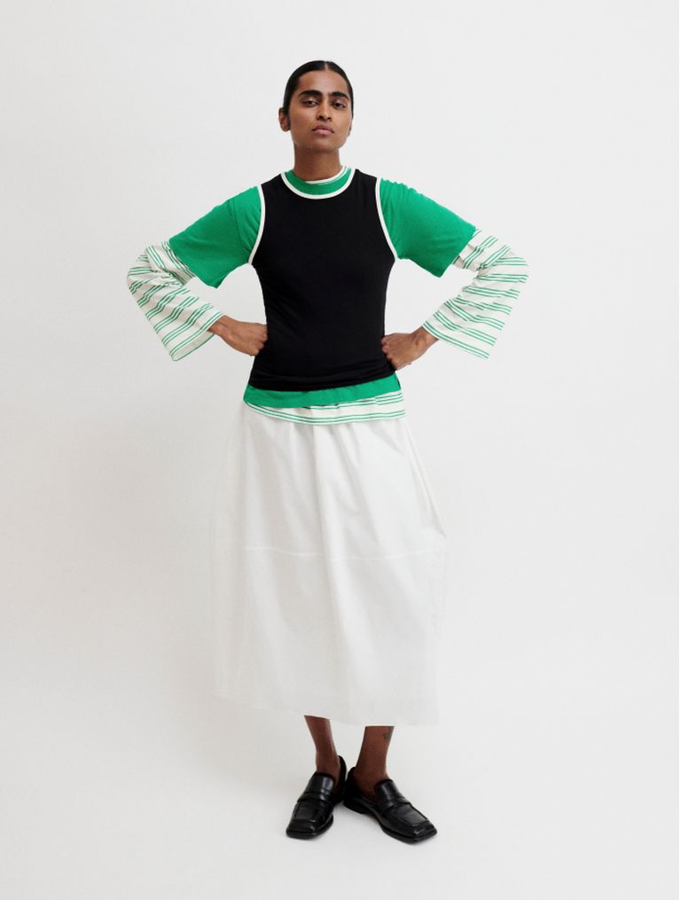 Person wearing a layered outfit with a black top over a green long-sleeve shirt, a white skirt, and black shoes, standing with hands on hips.