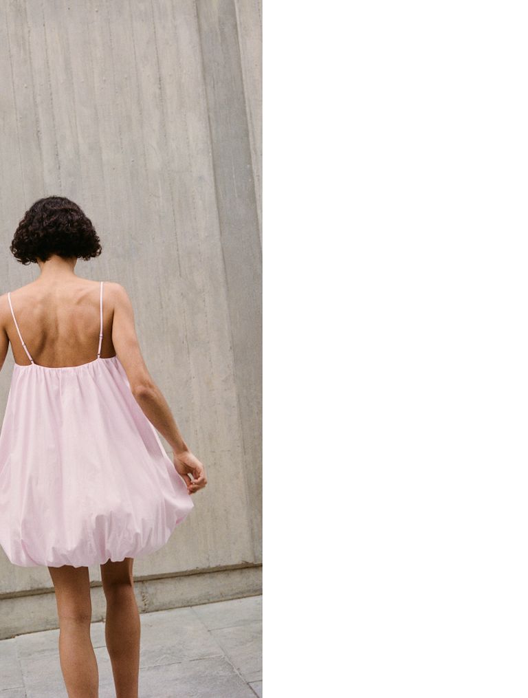 Person in a pink dress facing a concrete wall, with a bob hairstyle and thin straps, holding the hem of the dress.