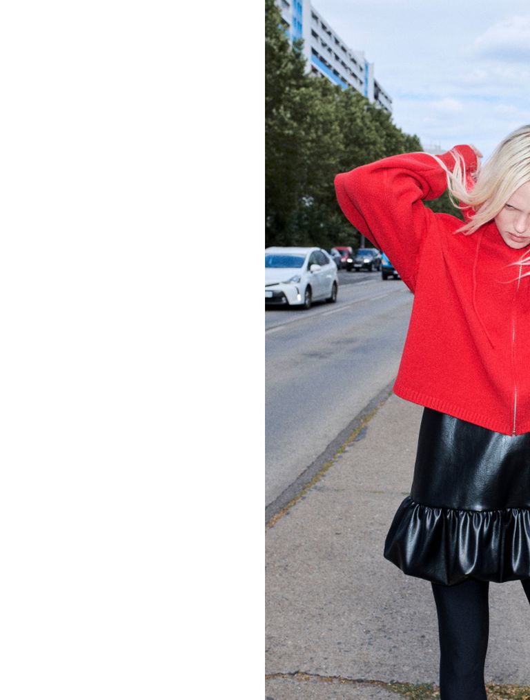 A person in a red sweater and black skirt stands on a street, adjusting their hair. Cars and trees are in the background.