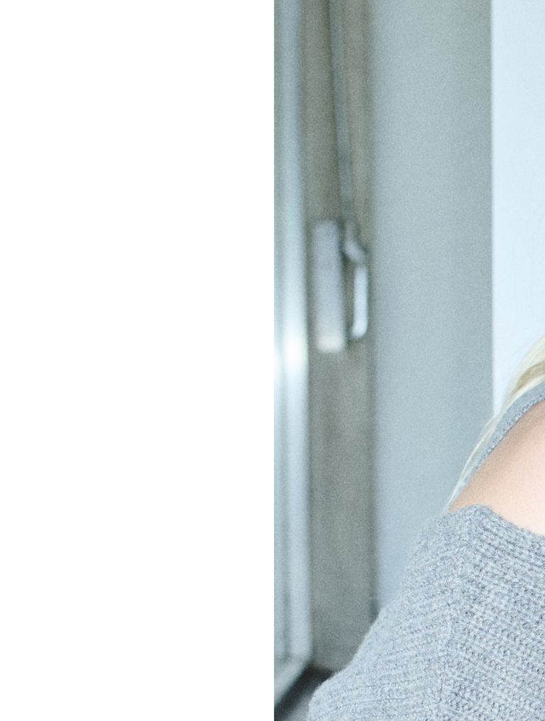 Blurred image showing part of a person in a gray sweater against an indoor background with a doorway and white wall.