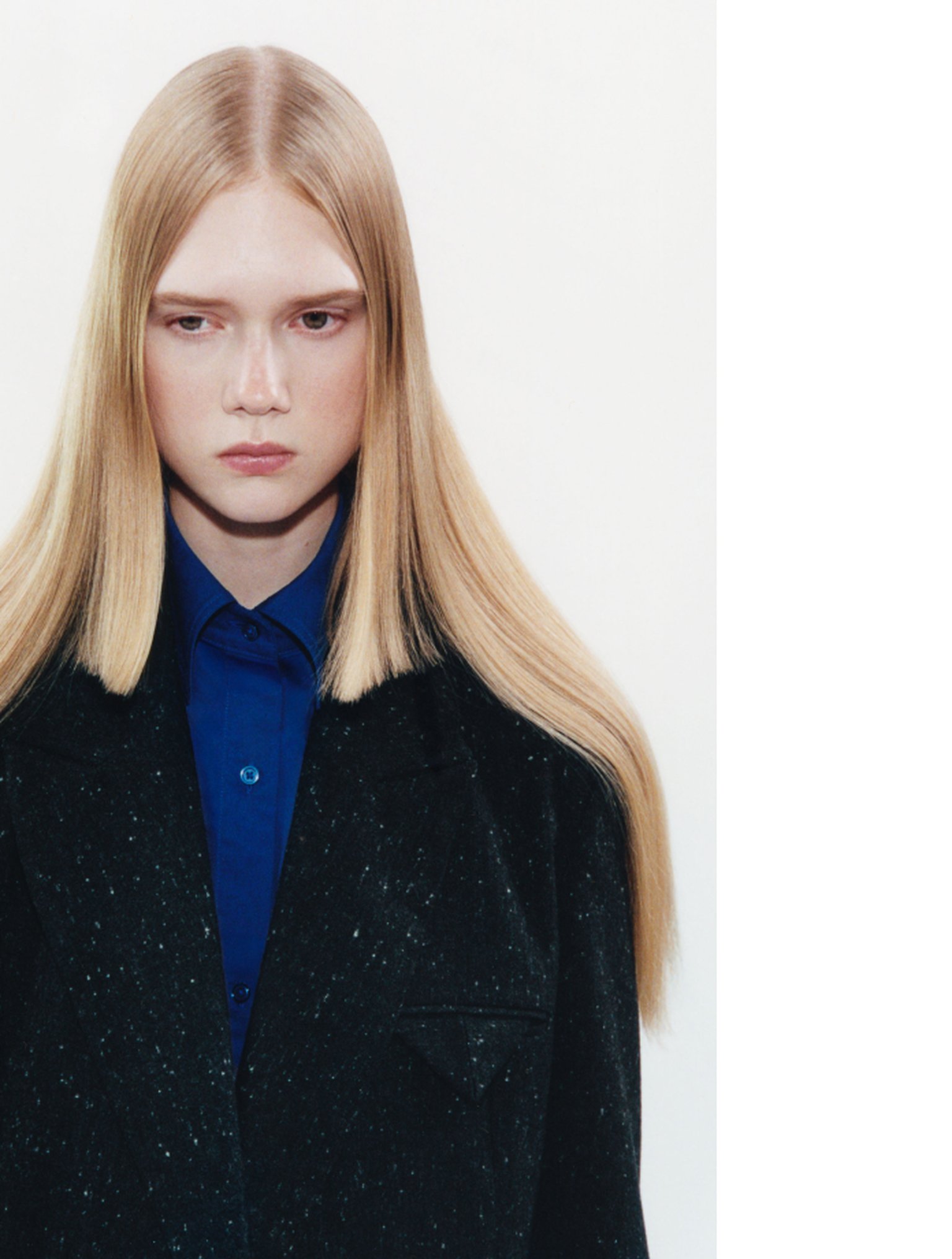 Person with long blonde hair wearing a blue shirt and dark coat, looking serious against a plain white background.