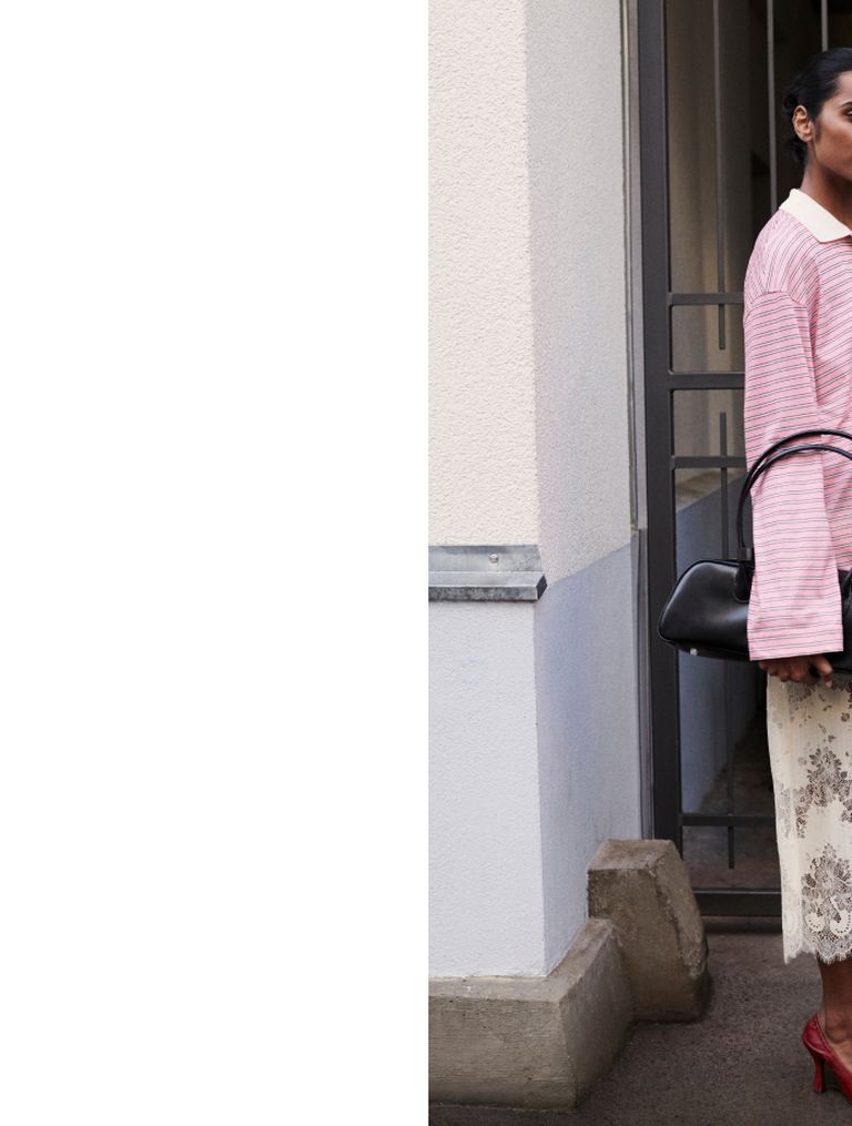 Person in a pink striped sweater and floral skirt, holding a black handbag, stands near a doorway, partially visible against a plain wall.