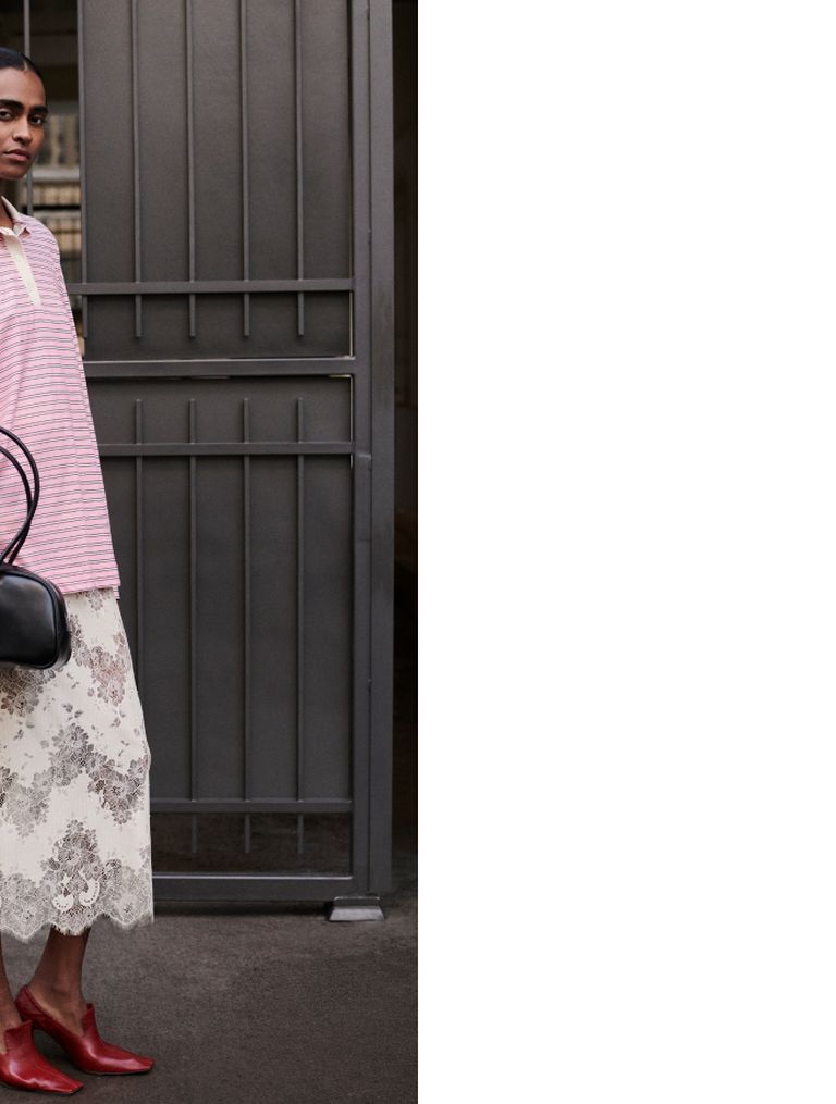 A person in a pink striped top and lace skirt stands near a metal gate, holding a black handbag. Red shoes complete the outfit.