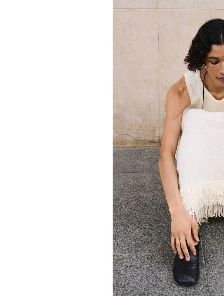 Person in a sleeveless white top with fringes sits on a gray stone floor, head down, with one hand on a black shoe.