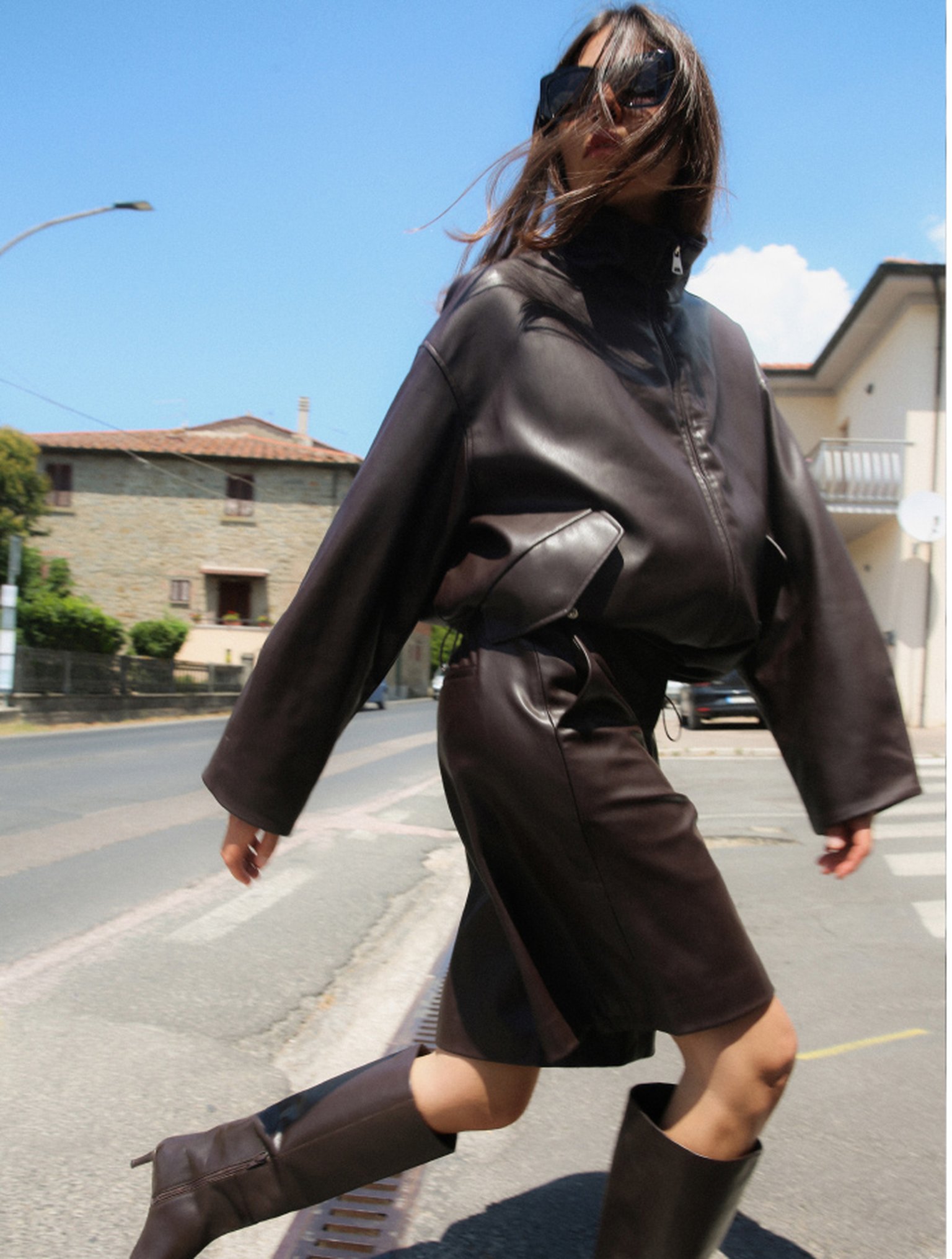 Woman in oversized black leather jacket and skirt, walking confidently on a sunny street, wearing sunglasses and knee-high boots.
