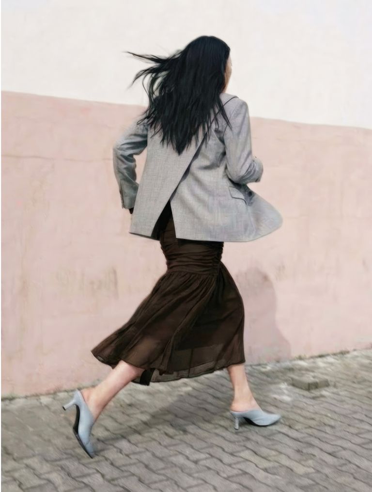 A woman with long black hair walks briskly in a gray blazer, brown skirt, and blue heels on a cobblestone path against a pale pink wall.