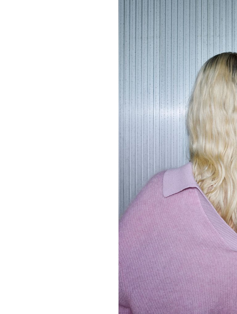 A person with long, wavy blonde hair wearing a pink sweater faces away from the camera, standing in front of a vertically striped wall.