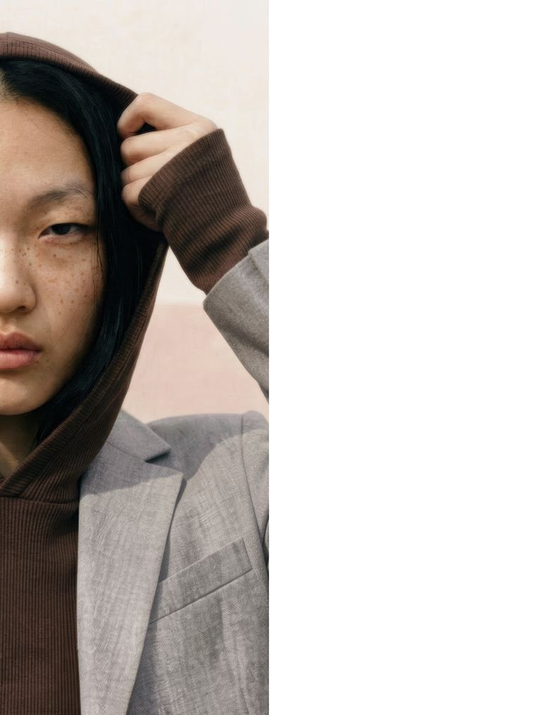 A person wearing a brown hoodie under a light gray blazer, partially visible, holding the hood with one hand against a light background.