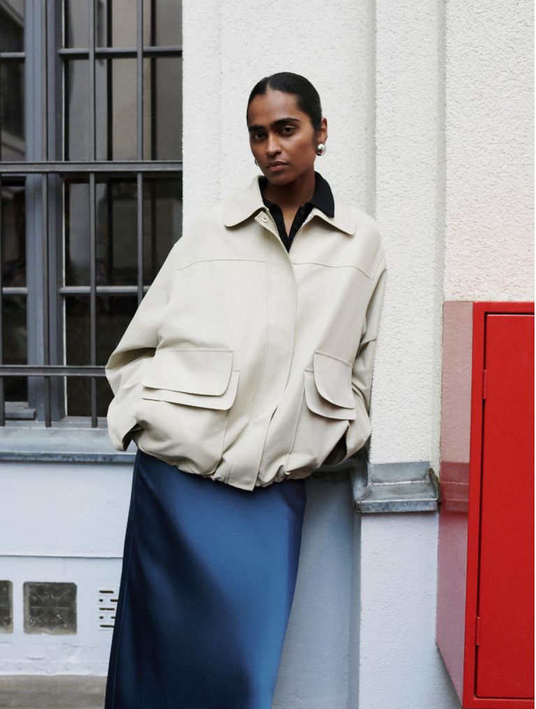 Person in a beige jacket and blue skirt leans against a building, near a red box, with a neutral expression.