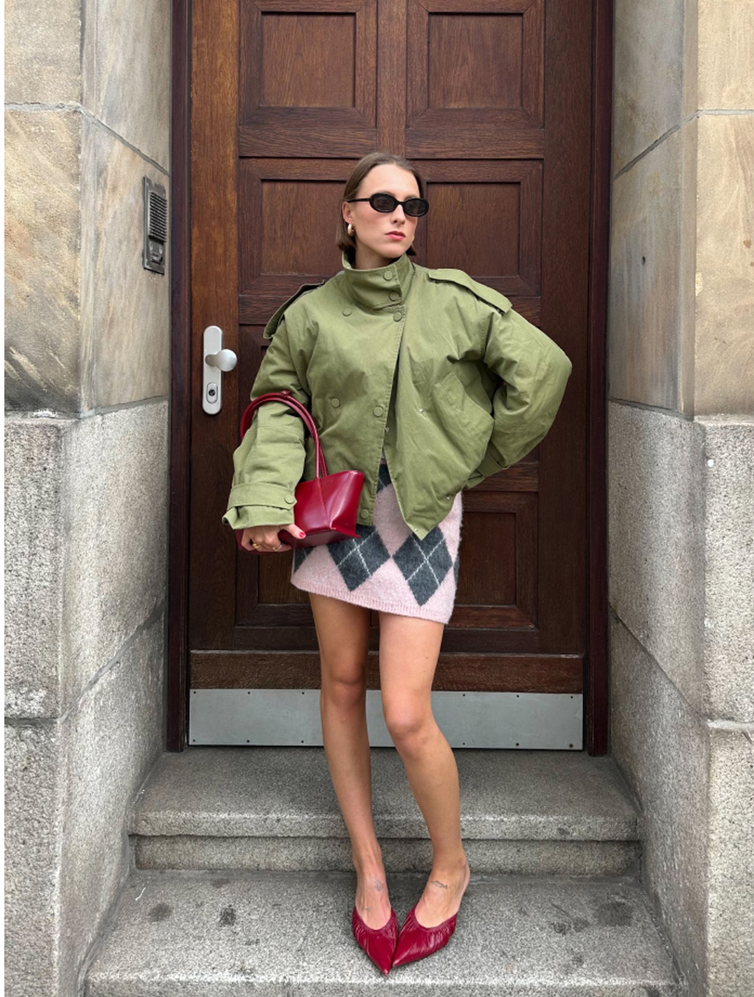 Woman in green jacket, pink argyle skirt, and red heels stands on steps, holding a red bag. Wooden door and stone walls in the background.