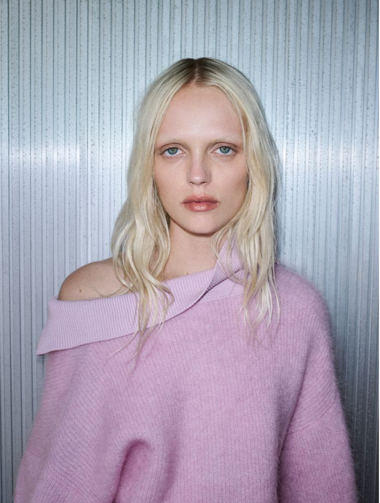 A person with long blonde hair wearing an off-shoulder pink sweater stands against a textured gray background, looking directly ahead.