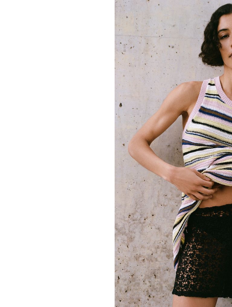 Person wearing a colorful striped tank top and black lace skirt, with one arm raised, stands against a textured concrete wall.