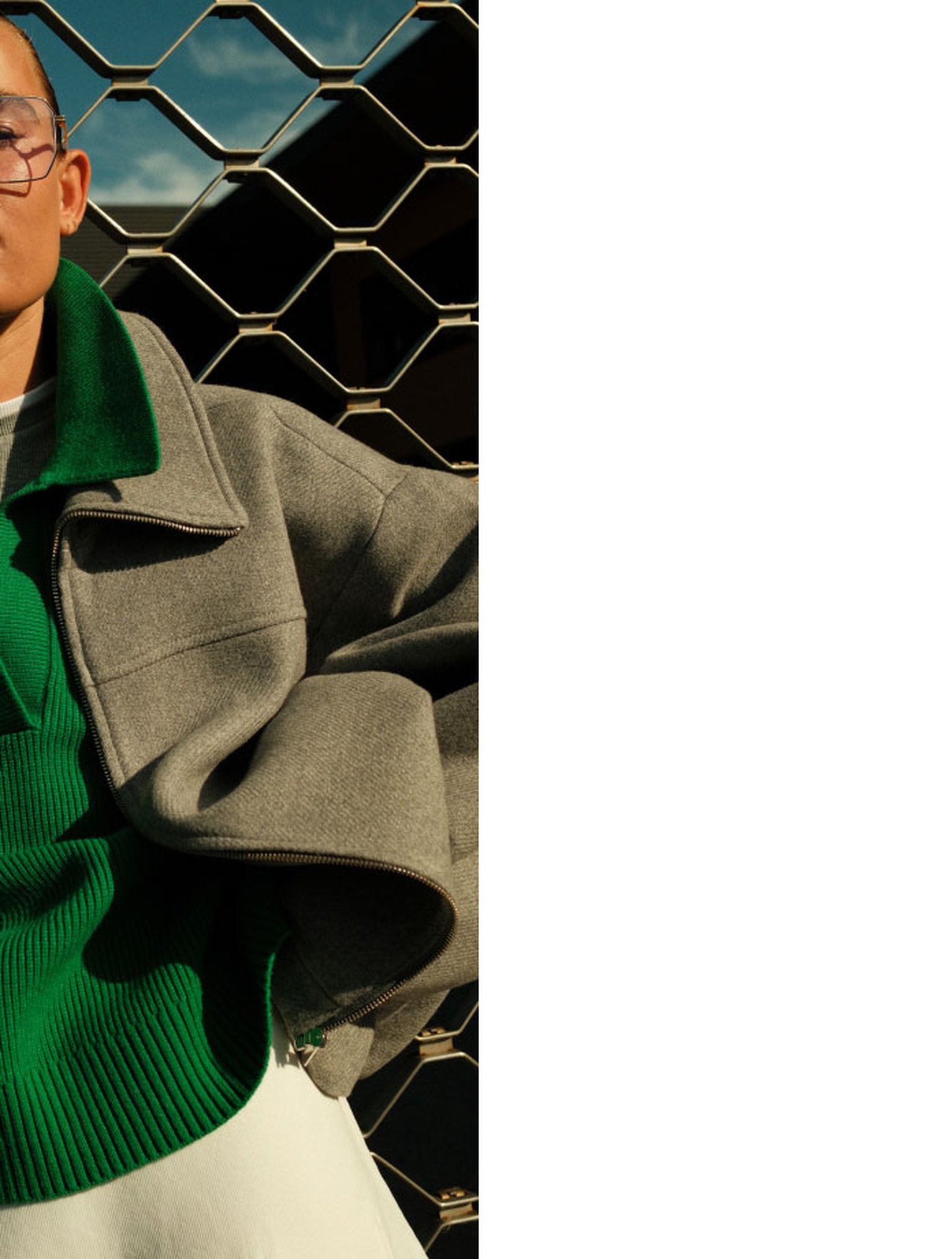 Person in a gray coat and green sweater stands in front of a metal fence, with part of their face and shoulders visible, against a partly cloudy sky.