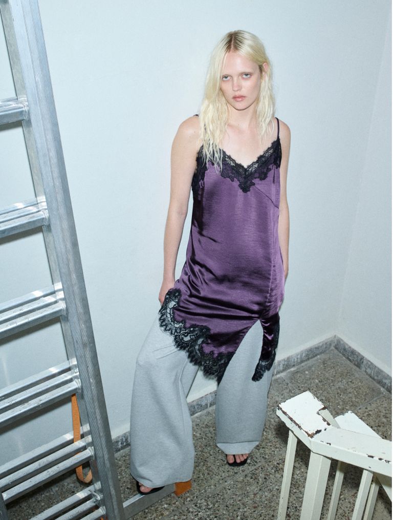 A person with long blonde hair wearing a purple satin top with black lace and gray pants stands near a metal ladder in a room with tiled flooring.