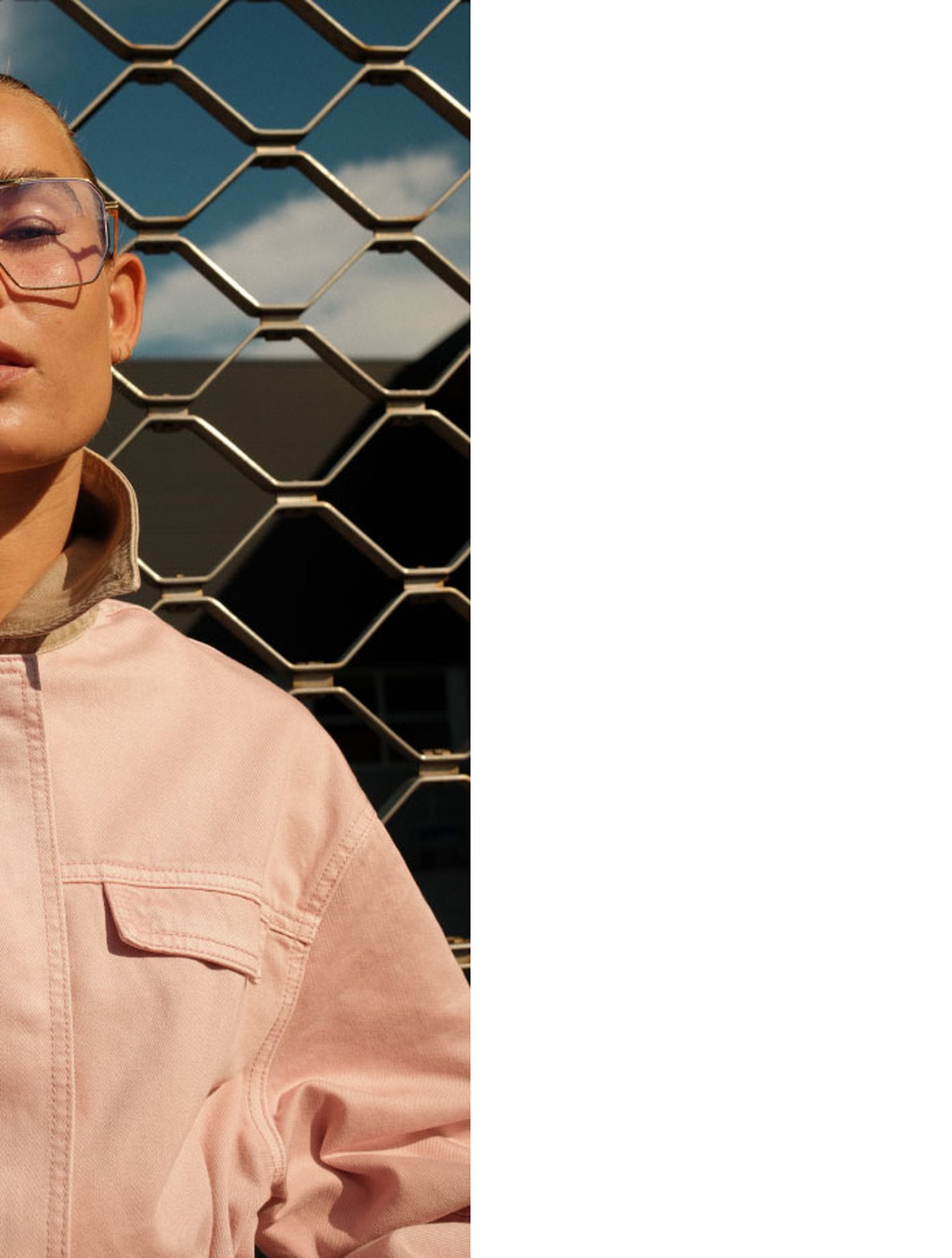 Person wearing transparent glasses and a light pink jacket, standing in front of a chain-link fence with blue sky and clouds in the background.