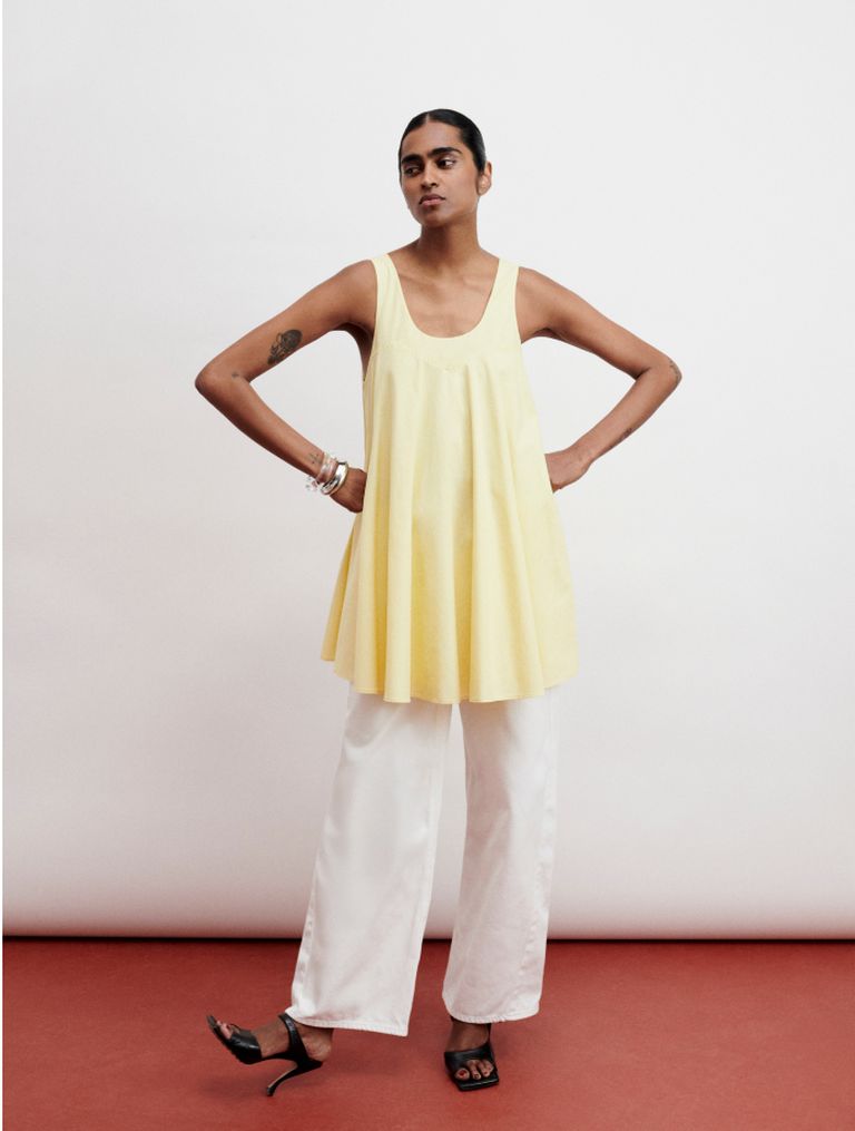 Person in a yellow sleeveless top and white pants, standing confidently on a red floor with hands on hips.