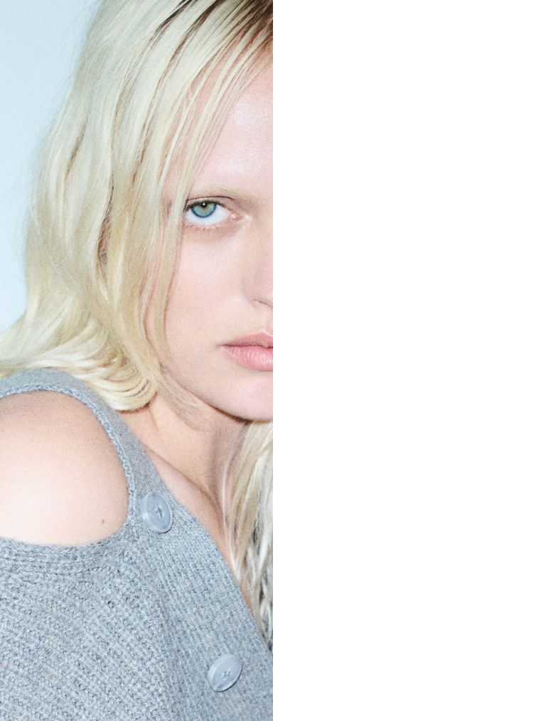 Close-up of a person with light hair and blue eyes, partially in view, wearing a grey sweater with buttons, against a light background.