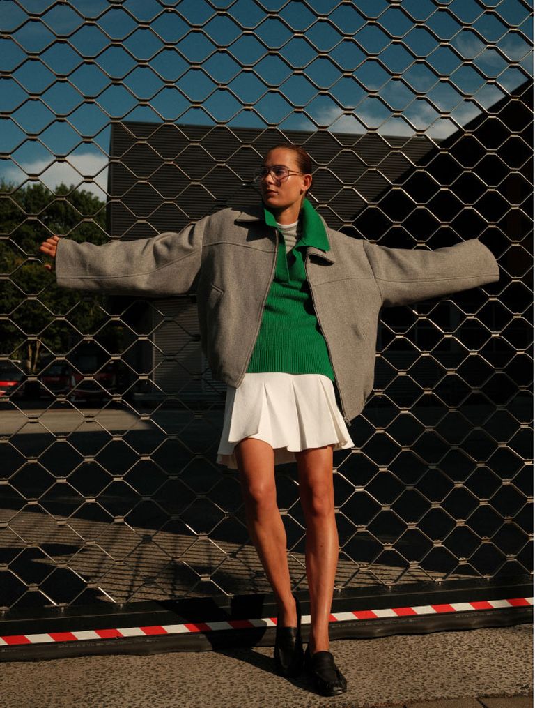 Person in a grey coat, green sweater, and white skirt stands with arms outstretched against a wire fence on a sunny day.