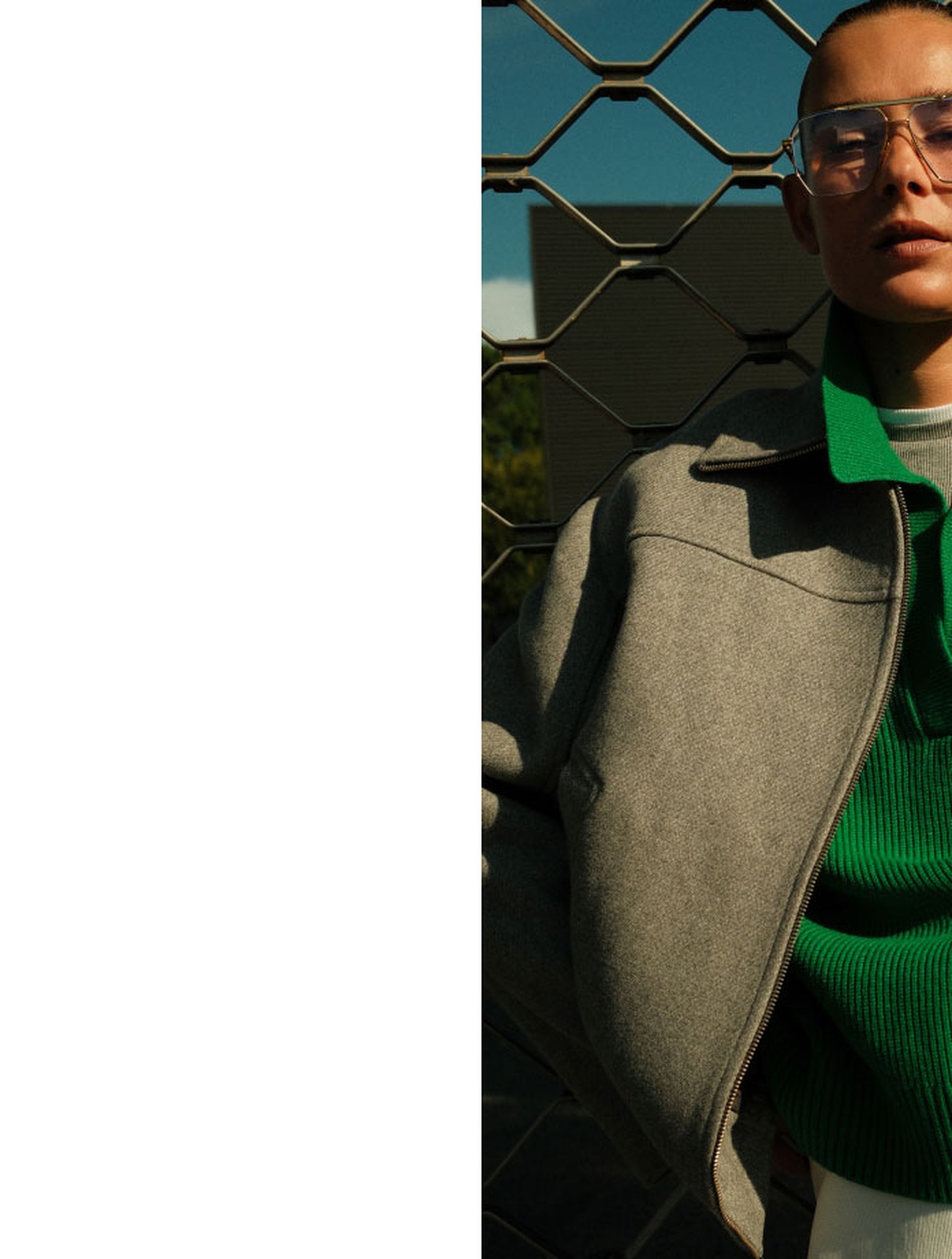 Person in a gray coat and green sweater stands against a wire fence, with sunglasses reflecting light.