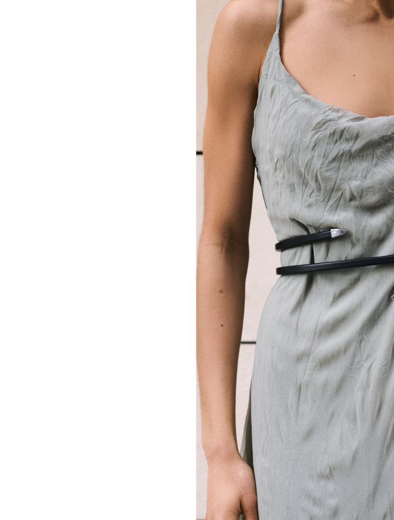 Person in a crinkled gray dress with thin straps and black belt, standing against a plain background.