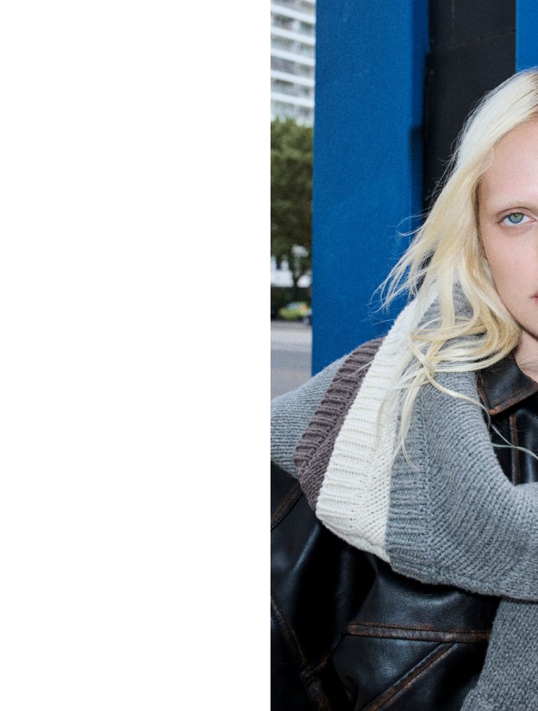 Person with blonde hair wearing a gray sweater and leather jacket, set against a blue wall and urban background.