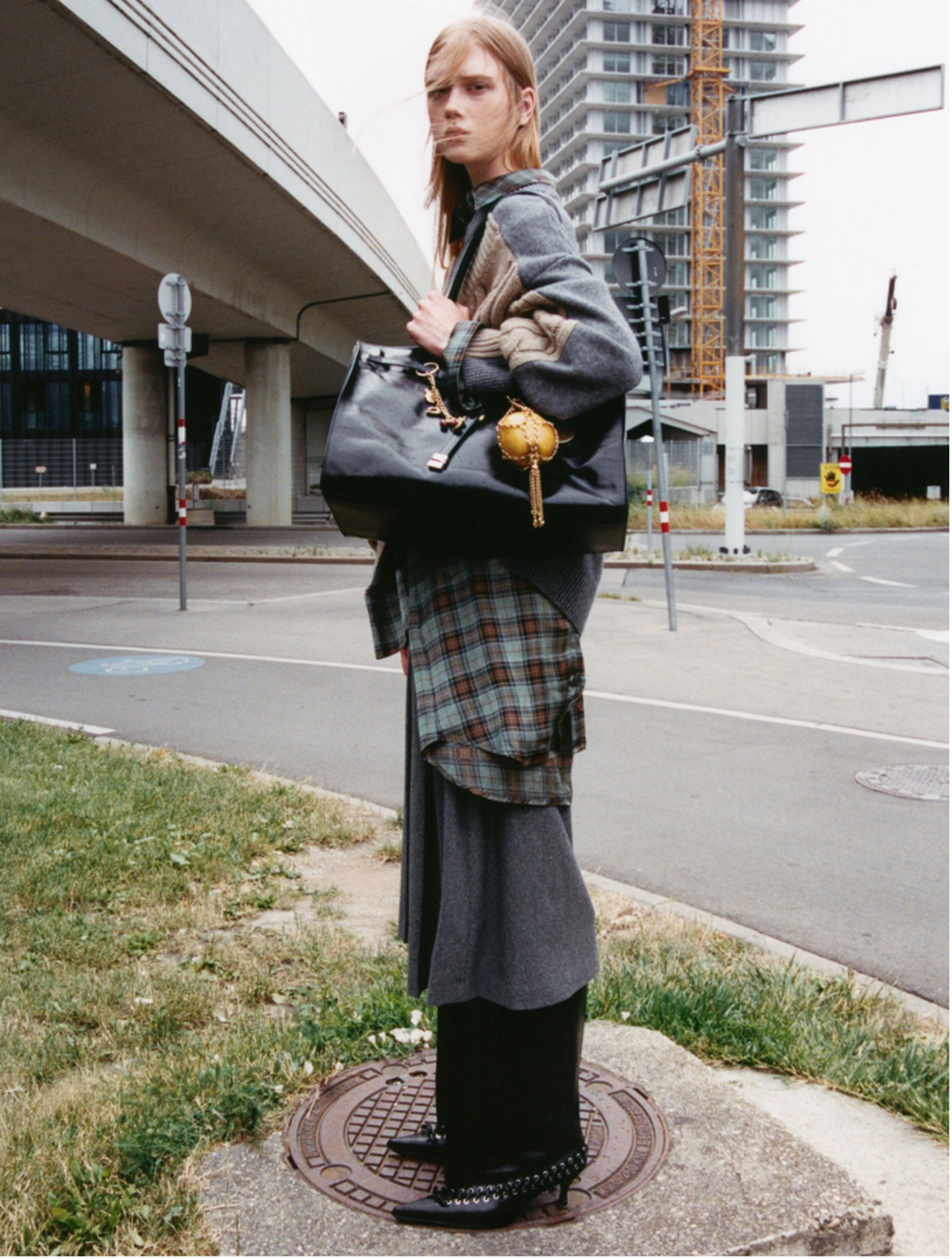 Person standing on a manhole cover in an urban setting, wearing layered clothing and holding a large black bag, with buildings in the background.