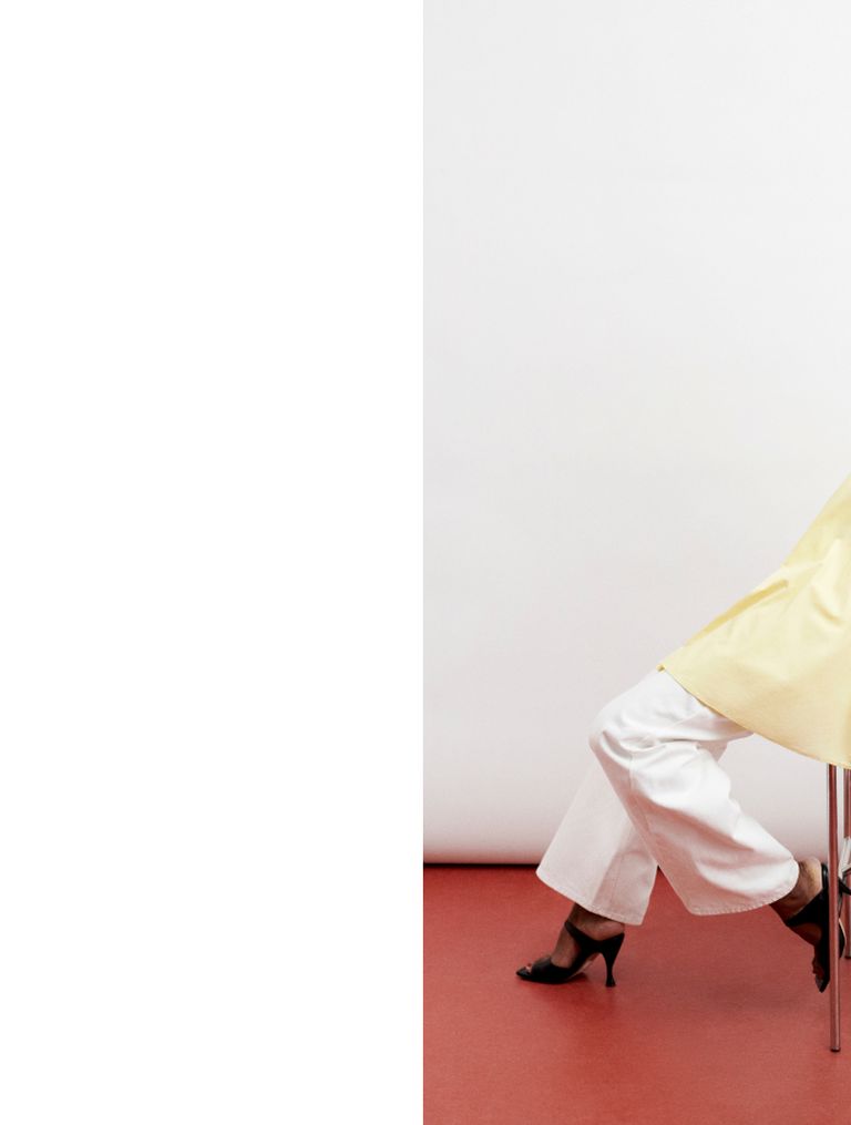 Person in yellow top and white pants seated on a stool against a two-tone red and white background. Legs and part of torso are visible.