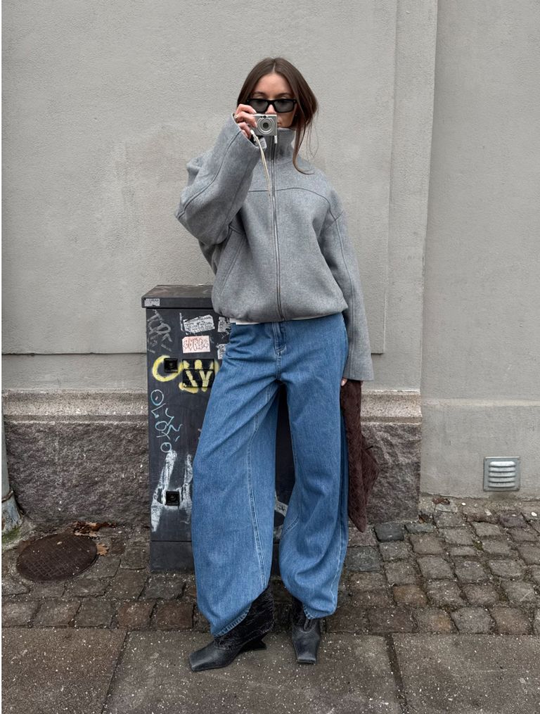 Person in oversized gray sweater and blue jeans takes a photo with a camera, standing on a cobblestone sidewalk beside a graffiti-covered utility box.