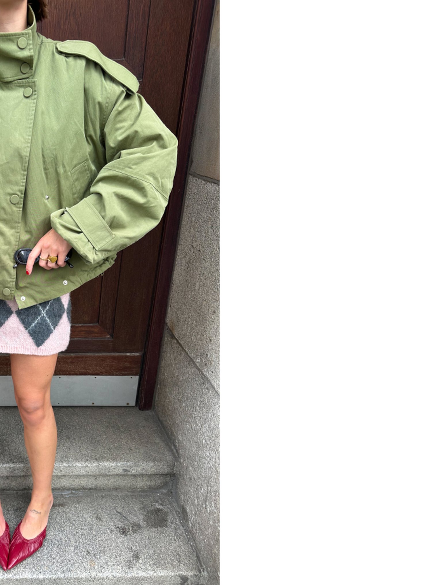 Person in a green oversized jacket and pink skirt stands on steps, holding a phone. Red shoes add a pop of color.