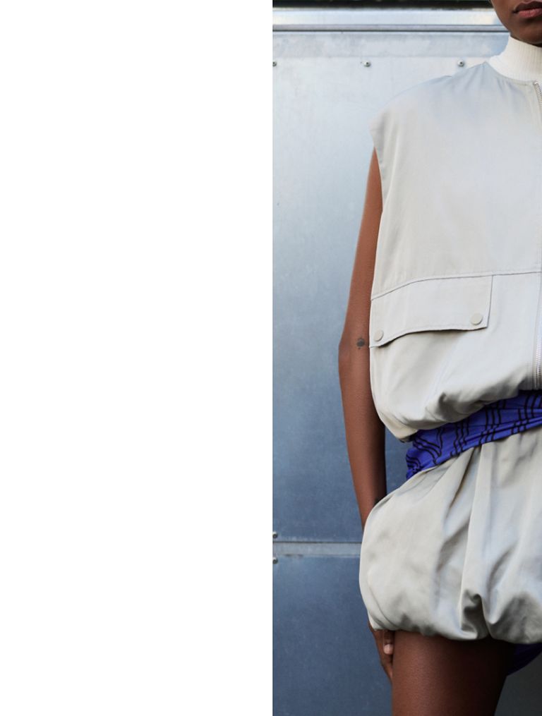 Person in a sleeveless, light gray outfit with a high neck and a blue belt, standing against a metallic background.