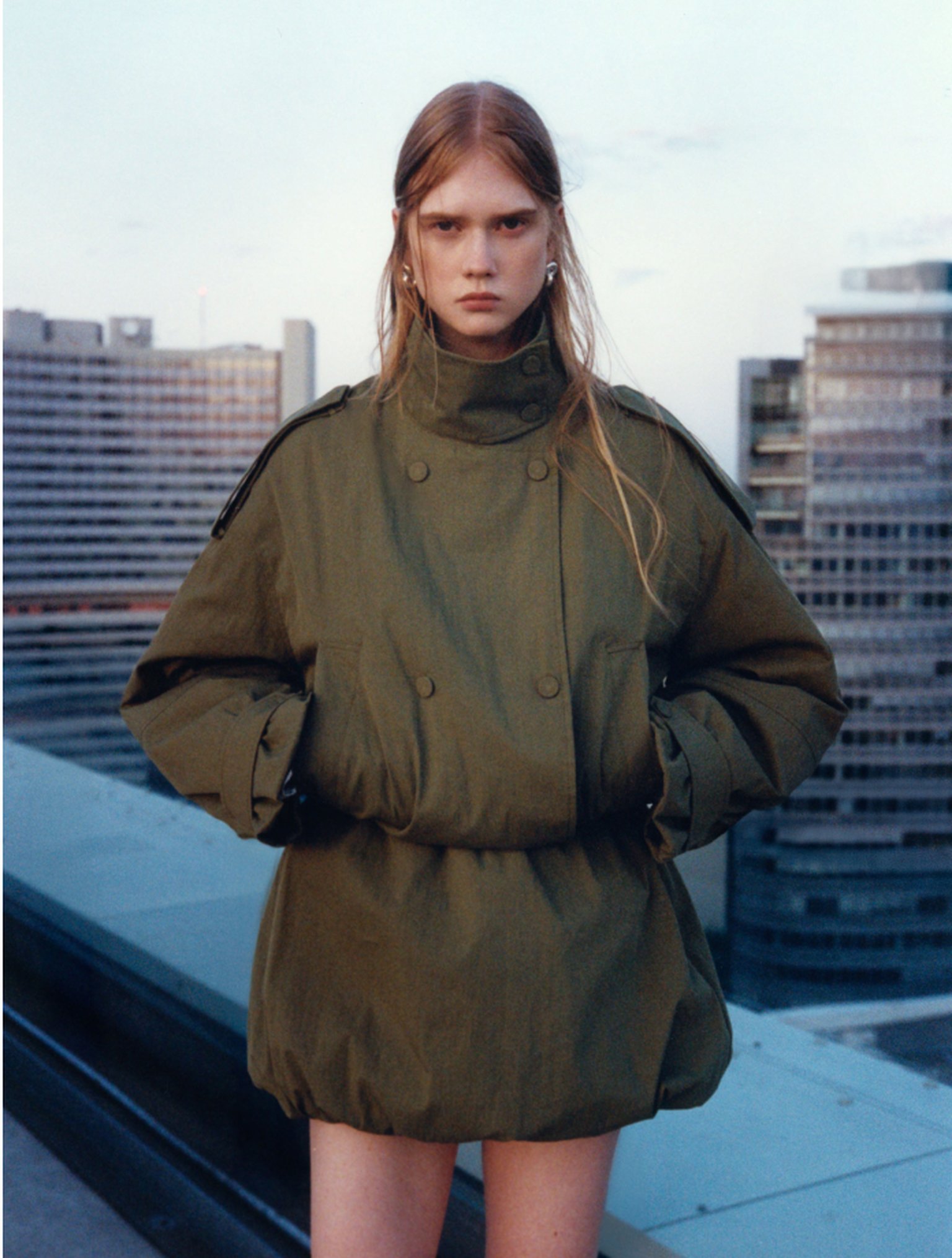 A person in a green oversized coat stands on a rooftop with modern buildings in the background at dusk.