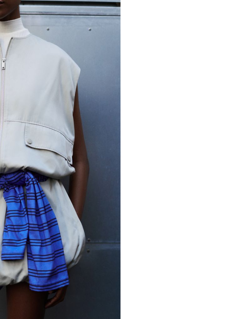 Person wearing a sleeveless beige vest and blue striped belt, standing against a metallic gray background.