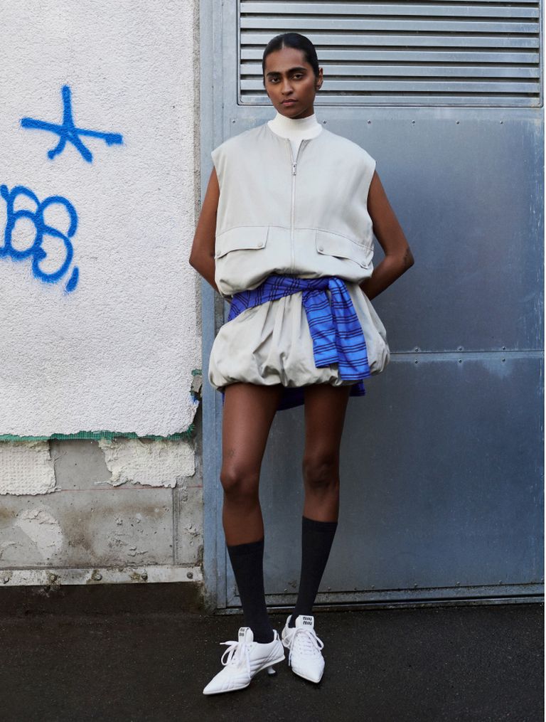 A person in a stylish outfit with a puffy skirt, vest, and knee-high socks stands against a wall with graffiti and a metal door.