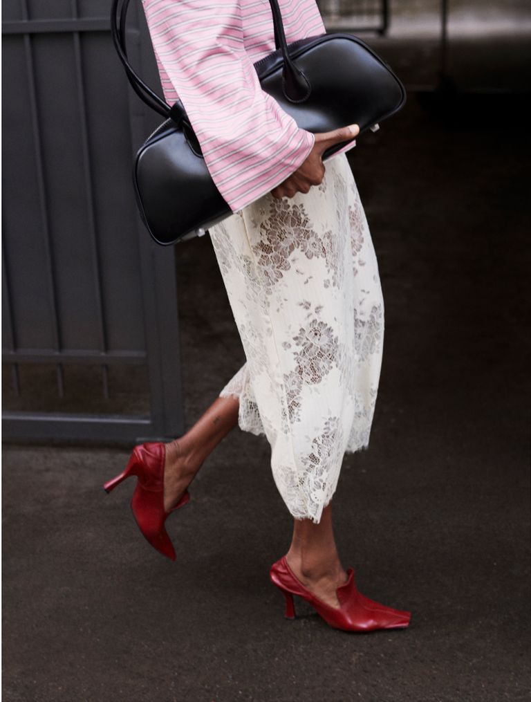 Person in pink striped shirt, white lace skirt, and red heeled shoes, carrying a black handbag, walking on a dark surface.
