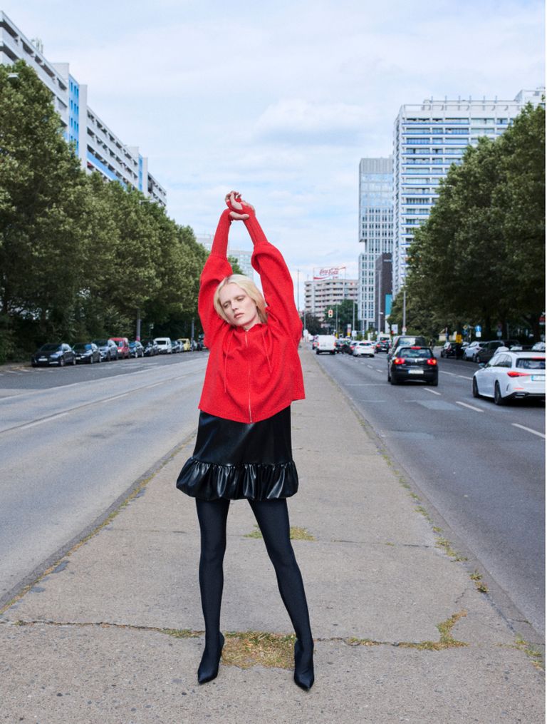 A person in a red sweater and black skirt stretches on a city sidewalk, with cars and tall buildings in the background.