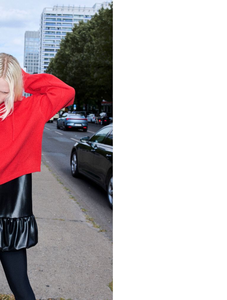 Person in a red sweater and black skirt stands on a city sidewalk, with cars and buildings in the background.