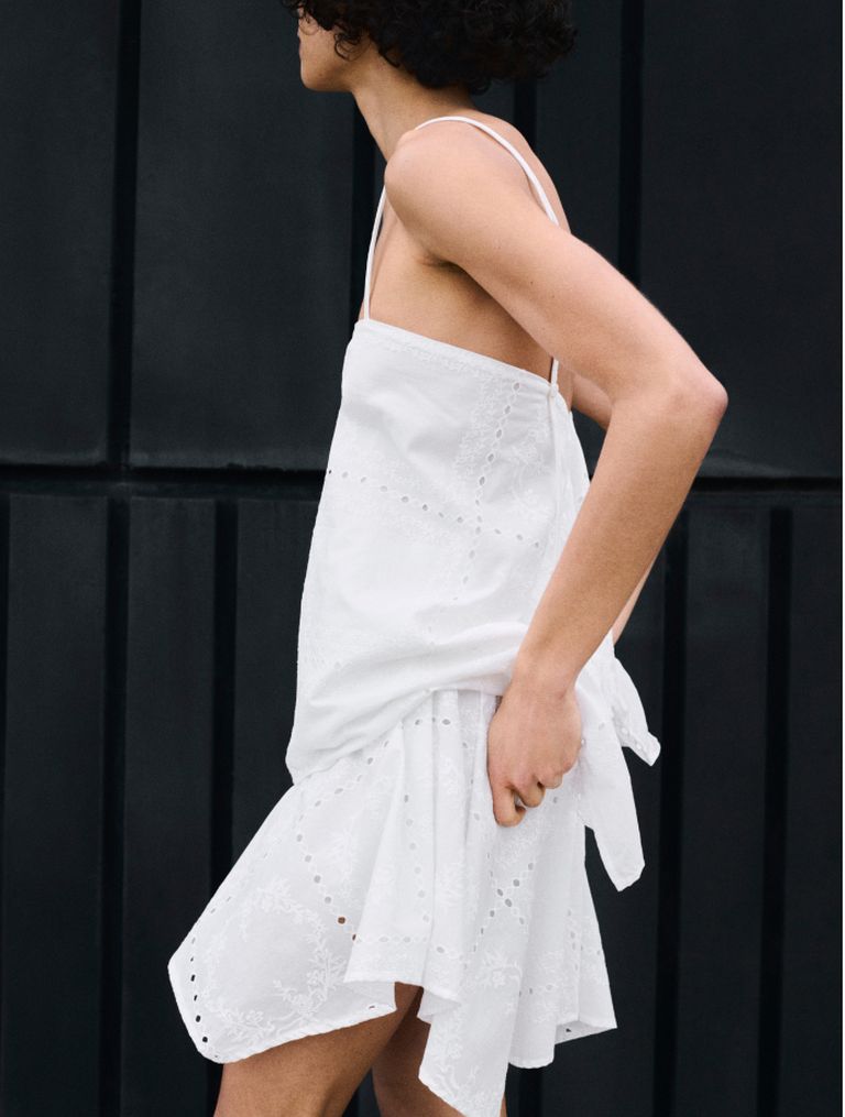 Person in a white, sleeveless dress with delicate eyelet pattern, walking against a dark background.