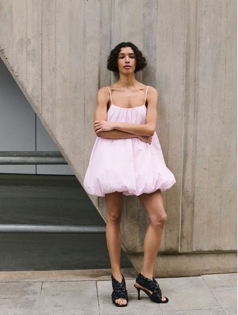 Person in a light pink bubble dress and black heels stands confidently against a modern concrete wall backdrop.