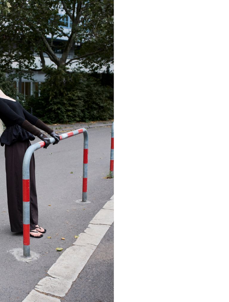 Person stretching against a red and white barrier on a paved path, surrounded by greenery. Upper body extends out of frame on right.