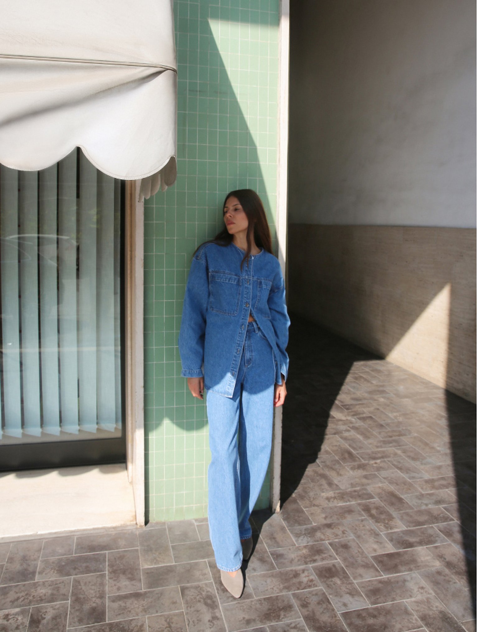 Woman in double denim stands against a green tiled wall, partially in shadow, on a tiled floor under an awning.