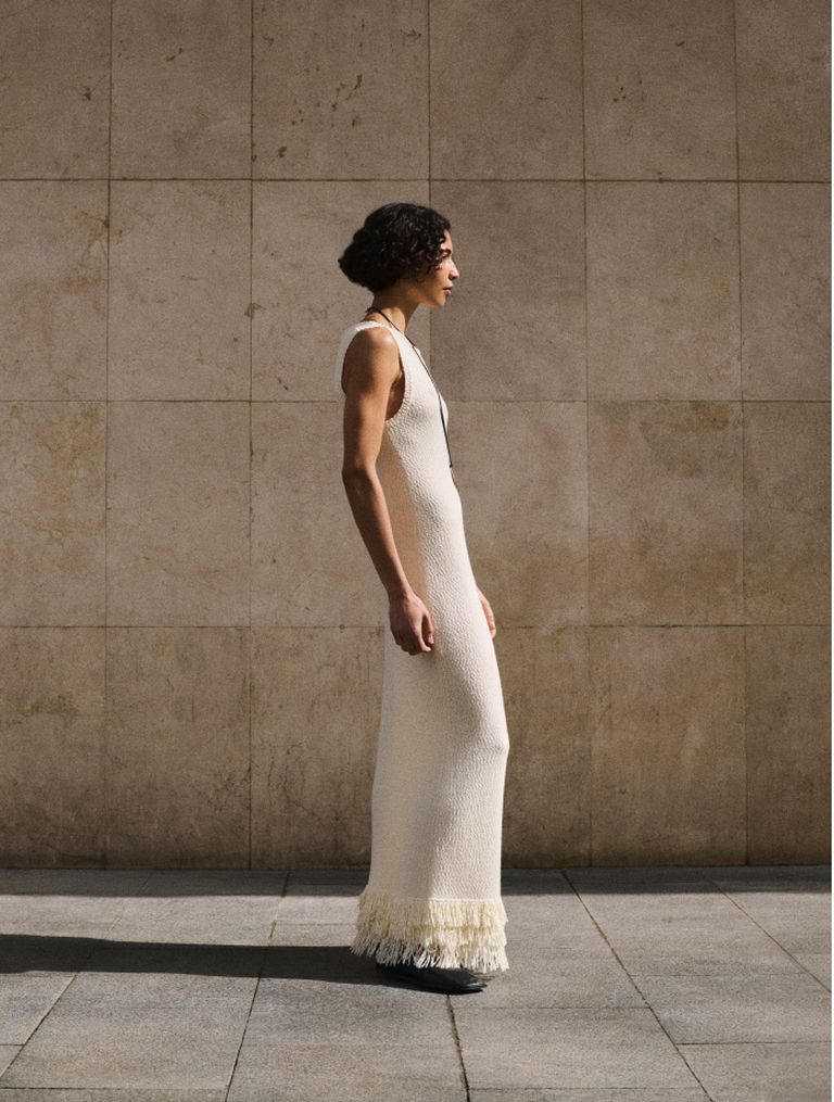 Person in a sleeveless, cream-colored dress with fringe hem, standing sideways against a textured stone wall.