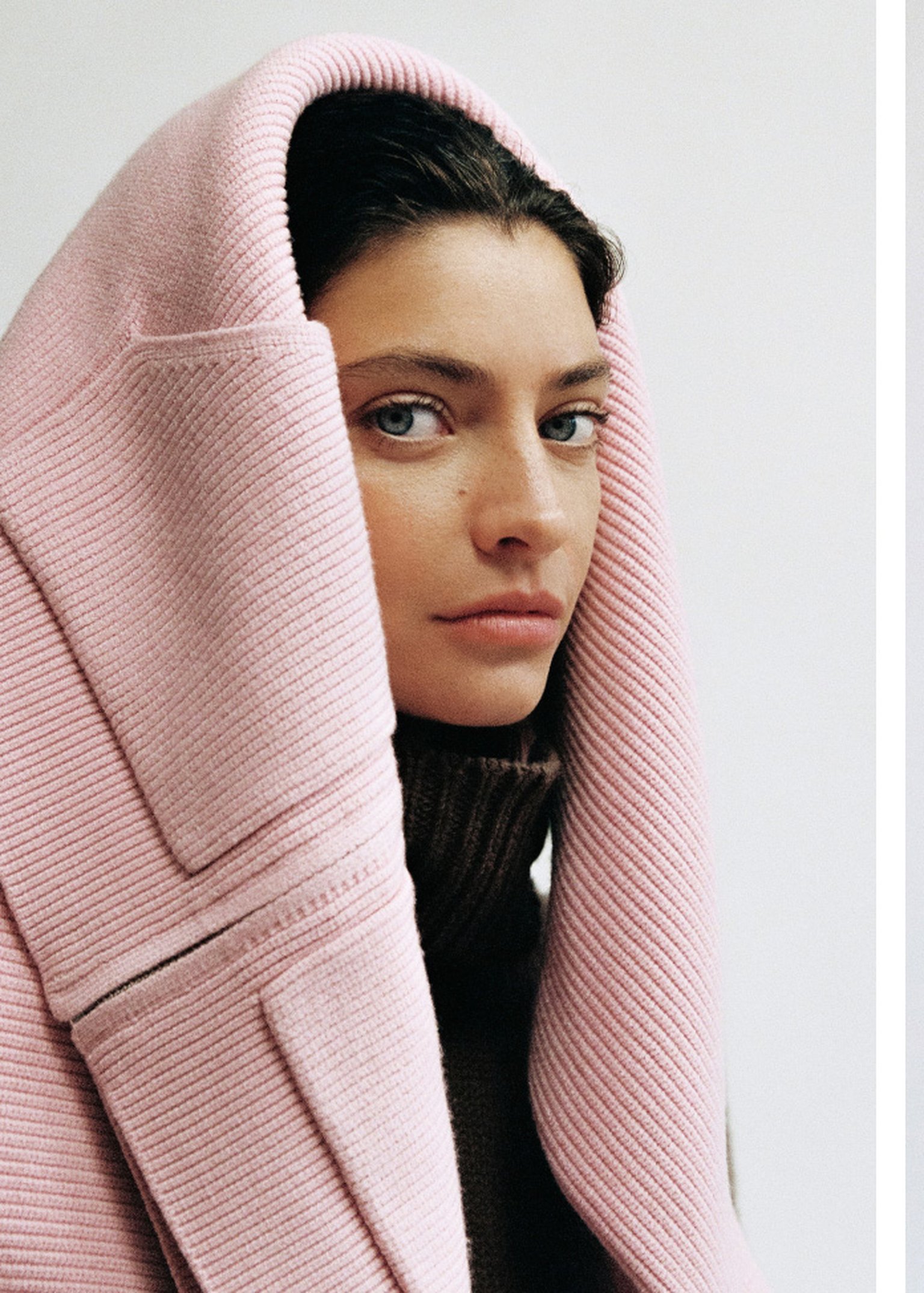 Woman wearing a pink ribbed coat with the hood up, over a brown sweater, gazing to the side against a neutral background.